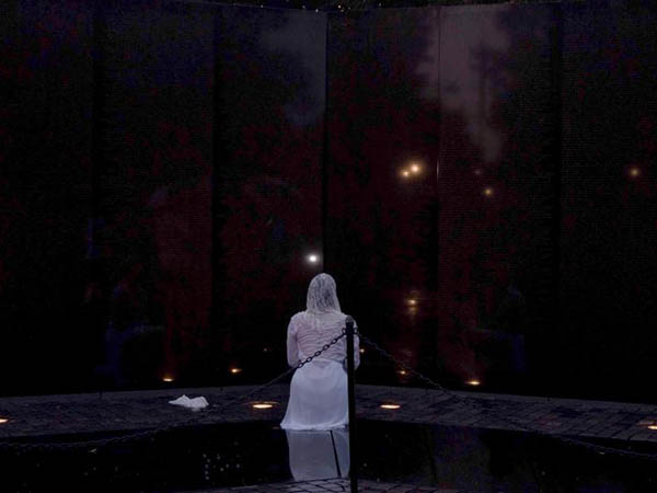 A woman in white kneels in front of a black sky with several lights twinkling in the distance